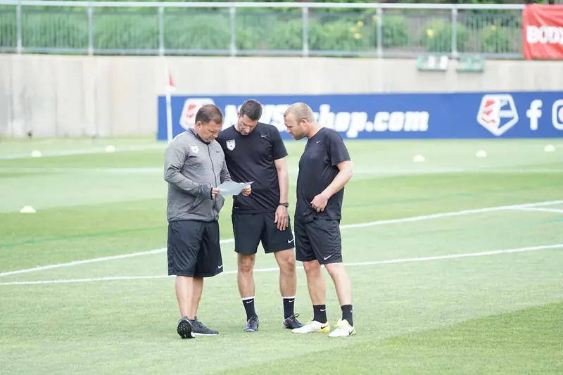Three coaches standing together on a pitch, looking over notes
